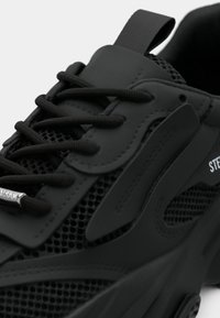 Scarpa sportiva nera con parte superiore in mesh strutturato, dettagli in sintetico liscio, lacci piatti e suola in gomma con dettagli di branding visibili.