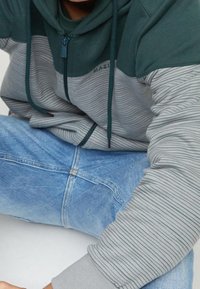 Osoba w zielono-szaro paskowanym hoodie na zamek i niebieskich dżinsach, siedząca z nogami skrzyżowanymi i ramionami opartymi na kolanach.