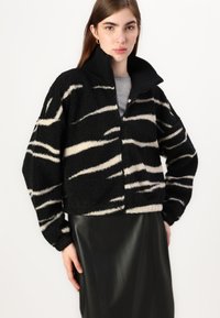 Svart og kremmønstret fleece-jakke med høy krage og glidelås, med en kort design og ribbestrikkede mansjetter.