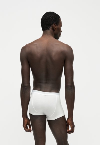 Άνδρας με σκουρόχρωμο δέρμα και κοντά σγουρά μαλλιά, που φοράει λευκά μπόξερ, στέκεται με την πλάτη προς την κάμερα μπροστά σε απλό λευκό φόντο.