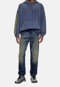 Hoodie en denim bleu avec une poche avant et un graphique jaune, associé à un jean tapered bleu clair. Présente des baskets métalliques.
