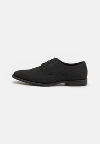Chaussure Oxford en suede noir avec une texture lisse, un bout arrondi et six œillets pour les lacets ; semelle en caoutchouc noir à profil bas avec des accents subtils.