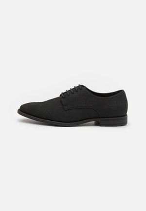Chaussure Oxford en suede noir avec une texture lisse, un bout arrondi et six œillets pour les lacets ; semelle en caoutchouc noir à profil bas avec des accents subtils.
