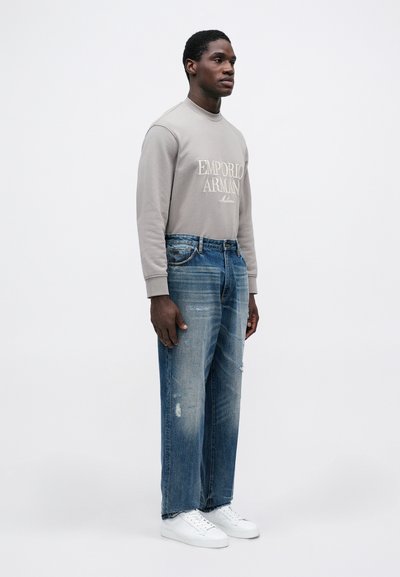 Camisa gris de manga larga con el logo de "Emporio Armani", jeans azules desgastados y zapatillas blancas. Diseño simple y corte relajado.