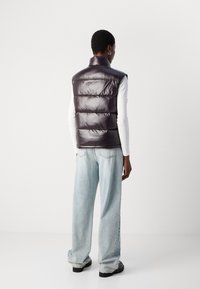 Osoba u sjajnom tamnom puffer prsluku, bijeloj majici dugih rukava, svijetloplavim širokim trapericama i crnim cipelama stoji okrenuta leđima.