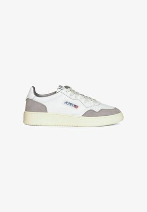 Baskets basses blanches avec des accents en daim gris, semelle crème, lacets, et un patch logo sur le côté avec un petit drapeau.