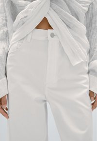 Vita byxor i bomullsblandning med raka ben, försedd med knappstängning, sidofickor och en slät yta. Matchas med en genomskinlig vit topp.