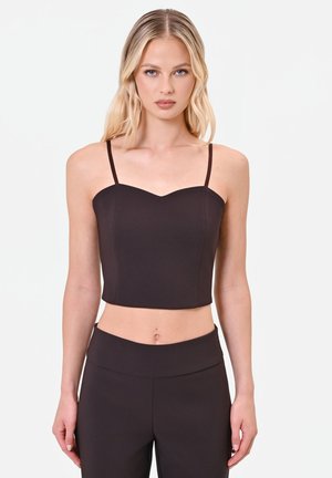 Młoda kobieta z długimi blond włosami, ubrana w czarny krótki top typu camisole oraz czarne spodnie z wysokim stanem, stojąca na jednolitym tle.