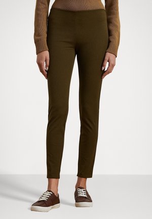 Persoon die olijfgroene aansluitende broek draagt, bruine sneakers met witte veters en een bruine gebreide top met lange mouwen, staand tegen een witte achtergrond.