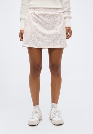 Άτομο που φοράει λευκή μίνι φούστα με μοτίβο ροζ φύλλων διάσπαρτο, λευκό μακρυμάνικο τοπ, λευκές κάλτσες και λευκά αθλητικά παπούτσια.