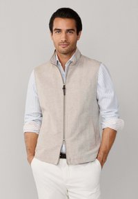 Homme portant une veste beige zippée sur une chemise à rayures bleues avec les manches retroussées, debout devant un fond gris clair uni.