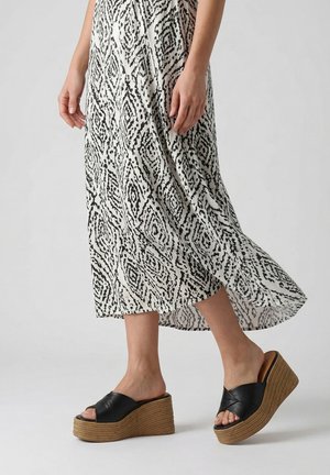 Žena v černých tkaných sandálech na klínku a splývavých bílých šatech s černým diamantovým vzorem kráčí na jednobarevném pozadí.