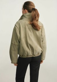 Przesadna oliwkowa zielona kurtka z wysokim kołnierzem, prążkowymi mankietami i ściąganym pasem, z teksturowanym, lekko zużytym materiałem.