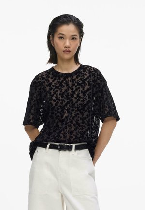 Mladá žena nosi priehľadný čierny top s kvetinovým vzorom, biele nohavice a čierny opasok, stojí s rukami v vreckách.