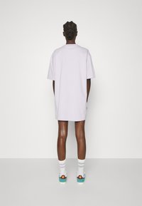 Ljuslila, oversized t-shirt med korta ärmar. Kombinerad med turkosa sneakers och vita, pastellrandiga strumpor. Bakifrån, enkel bakgrund.