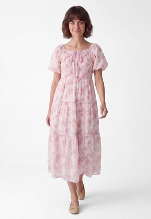 Kvinde med kort brunt hår iført en lyserød blomsterprintet midi-kjole med pufærmer og beige ballet flats, stående mod en hvid baggrund.