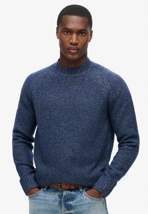 Suéter de punto azul marino con cuello redondo, puños y dobladillo acanalados; textura sutil y corte relajado. Combinado con jeans de mezclilla azul claro.