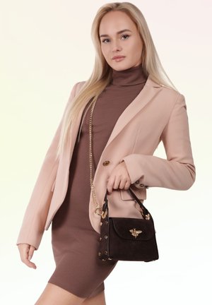 Blonde woman wearing brown dress and light pink blazer holding small black handbag with gold bee emblem, standing against light background.