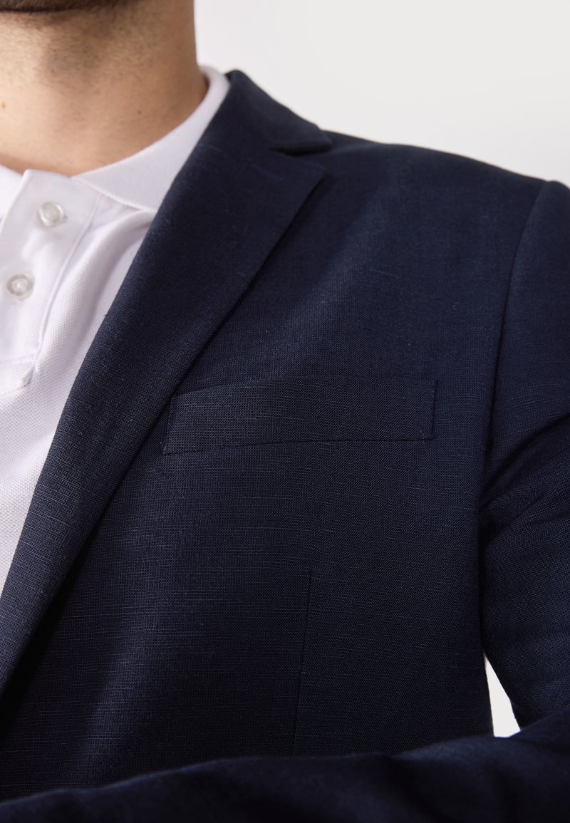 Blazer bleu marine en tissu texturé, avec une poche poitrine simple et des revers à cran, porté par-dessus un polo blanc à col boutonné.