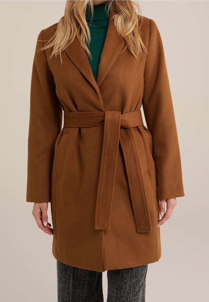 Bruine wollen mantel met ceintuur, een notchkraag, lange mouwen en twee zijzakken, gedragen over een groene coltrui, gecombineerd met grijze broek met patroon.