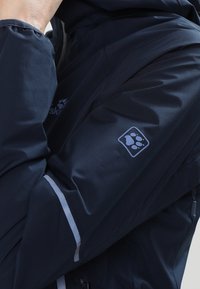 Personne portant une veste bleu marine avec un logo réfléchissant en forme d'empreinte de patte sur la manche et une bande réfléchissante près du coude.