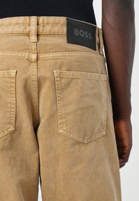 Pantaloni in denim beige con finitura testurizzata, caratterizzati da un'etichetta nera con il logo BOSS in vita e due tasche posteriori con bordi cuciti.