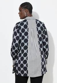 Chemise à pois noir et blanc avec des tailles de cercles variées. Manches longues, poignets boutons et un détail de pli dans le dos. Texture de tissu lisse.