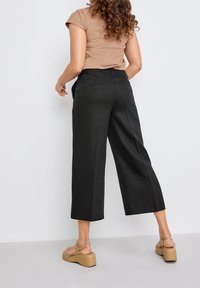 Svarta vida byxor med en slät textur, som har bakfickor och en midjehöjd som slutar i midjan. Modellen bär en figurnära brun topp och beige plattformssandaler.