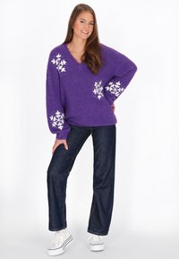 Maglione viola, morbido e lavorato a maglia con scollatura a V e motivi di fiocchi di neve bianchi; abbinato a jeans blu scuro e sneaker bianche.