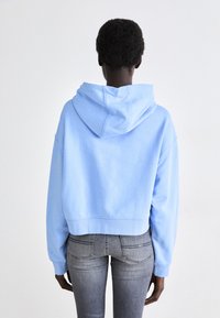 Haut bleu coupé en tissu doux, avec une capuche à cordon et des poignets côtelés. Porté avec un jean gris ajusté.