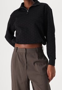 Sweat-shirt court noir à fermeture éclair avec motif de logo répété, associé à un pantalon marron taille haute, coupé sur mesure et à la texture lisse.