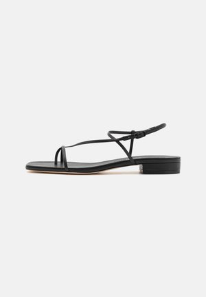 Sandale plate noire avec des fines brides qui se croisent sur le pied et une bride à la cheville avec une petite boucle.