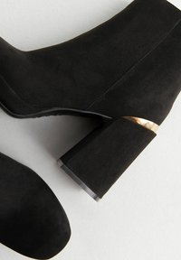 Bottines en suédine noire avec des talons blocs mettant en avant des détails en or, présentées sur une surface claire.