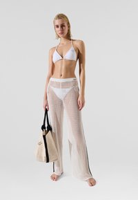 White bikini with triangular top, paired with sheer white netted pants featuring a striped accent. Model holds a beige tote bag.