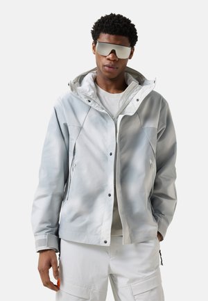 Mężczyzna w jasnoszarej kurtce z kapturem, białych spodniach i dużych lustrzanych okularach przeciwsłonecznych, stojący z jedną ręką w kieszeni.