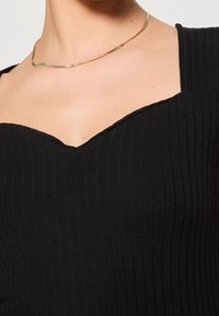 Gros plan d'une personne portant un haut noir côtelé avec un col carré et un fin collier en chaîne dorée.