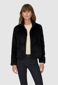 Schwarze Kunstpelzjacke mit hohem Kragen, im cropped Design und mit Seitentaschen, getragen über einem cremefarbenen Pullover und dunklen Jeans.