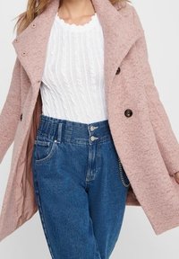 Cappotto in misto lana rosa chiaro con ampio colletto e grandi bottoni neri, sovrapposto a un top in maglina bianca a coste e jeans in denim blu a vita alta.