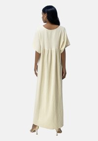 Robe longue de couleur crème en tissu texturé avec des manches courtes et larges et une coupe ample. Elle met en avant une taille froncée et une silhouette fluide.