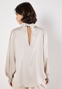 Blusa di seta beige con righe verticali, apertura a chiave sul retro e chiusura a fiocco. Presenta maniche a sbuffo e polsini arricciati per una maggiore texture.