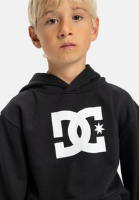 Čierna mikina s bielym logom so stylizovaným "DC" a hviezdou. Tkanina vyzerá mäkko, má štandardnú kapucňu a rebrové manžety.