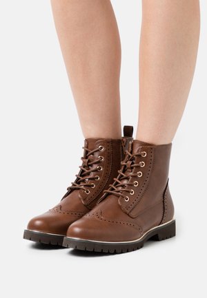 Bottines montantes en cuir marron à lacets avec détails brogue, portées sur des jambes nues sur un fond blanc.