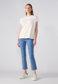 T-shirt blanc avec des rayures horizontales jaunes et un petit logo rouge, assorti à un jean skinny bleu et des baskets blanches. Design simple et décontracté.