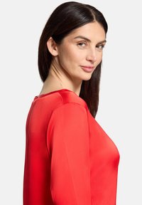 Rød satinbluse med rund halsudskæring, glat tekstur og lange ærmer. Stoffet har en subtil glans, der fremhæver dens levende farve.