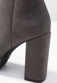 Harmaa korkeakorkoinen nilkkabooti, joka on valmistettu teksturoidusta materiaalista, varustettu paksulla neliön muotoisella korolla ja saumattomalla muotoilulla, jossa on hienovarainen sauma-yksityiskohta.