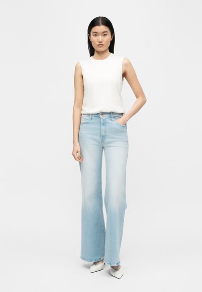 Γυναίκα με ίσια μαύρα μαλλιά, φορώντας λευκή αμάνικη μπλούζα, ανοιχτόχρωμα τζιν παντελόνια φαρδιάς γραμμής και λευκά μυτερά παπούτσια, στεκόμενη με το ένα χέρι στην τσέπη.