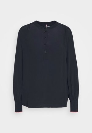 Blouse noire à manches longues avec col rond, patte de boutonnage à l'avant et poignets légèrement froncés avec bordure rouge et blanche.