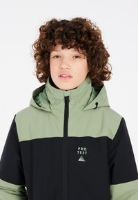 Veste à capuche noire et verte avec fermeture éclair à l'avant, arborant un logo sur la poitrine et une finition texturée. Conçue pour les activités de plein air.