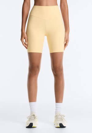 Femme portant un short cycliste taille haute jaune pâle associé à des chaussettes blanches et des chaussures de sport beiges, debout devant un fond uni.