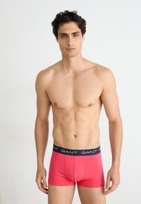 Rote Baumwollboxershorts mit navyfarbenem Bund, der mit weißem "GANT"-Branding versehen ist, im figurbetonten Design und mit weicher Textur, präsentiert an einem männlichen Modell.